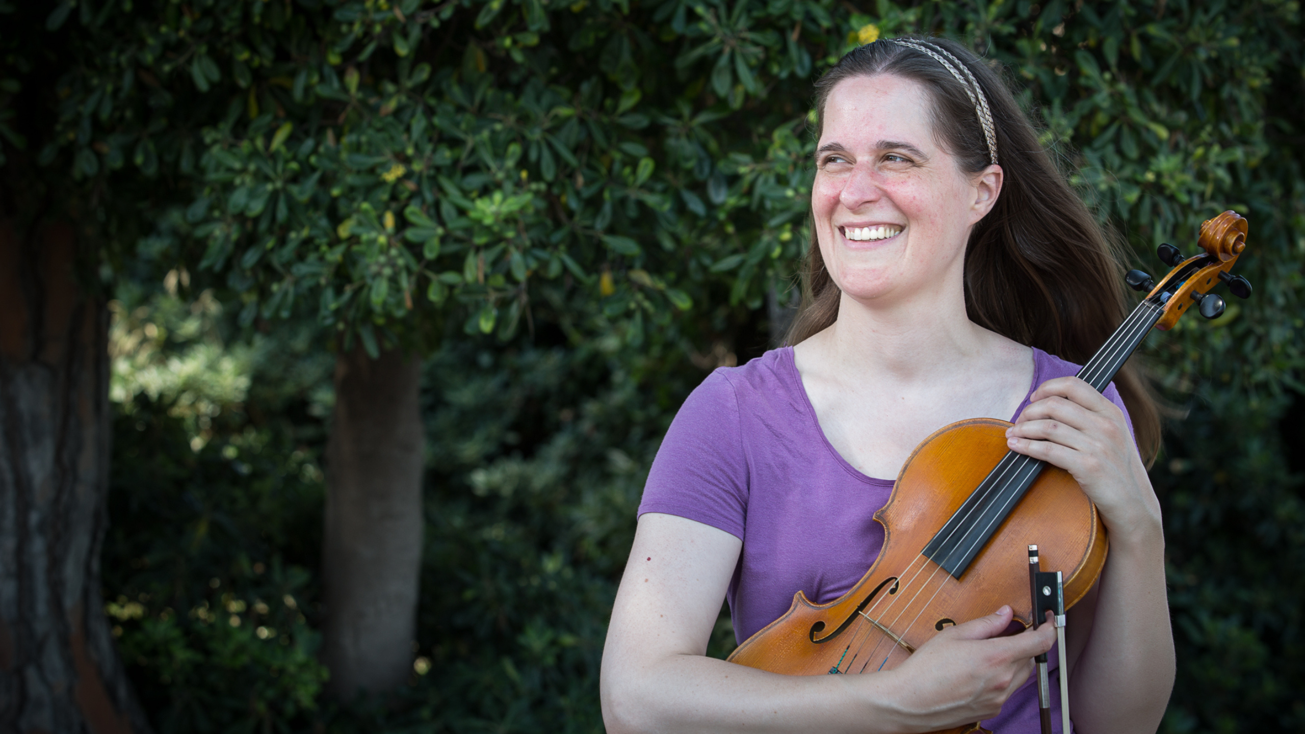 Leah Irby holding her viola, wearing a purple top, with trees in the background