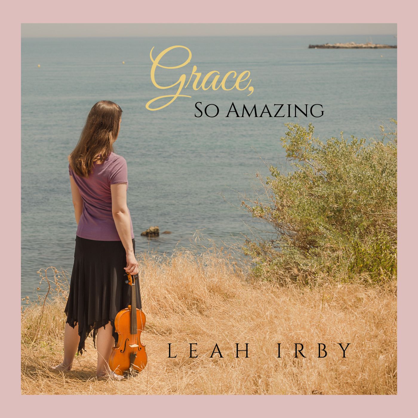 Leah Irby standing in grass overlooking the ocean, looking into the sea while holding her viola.