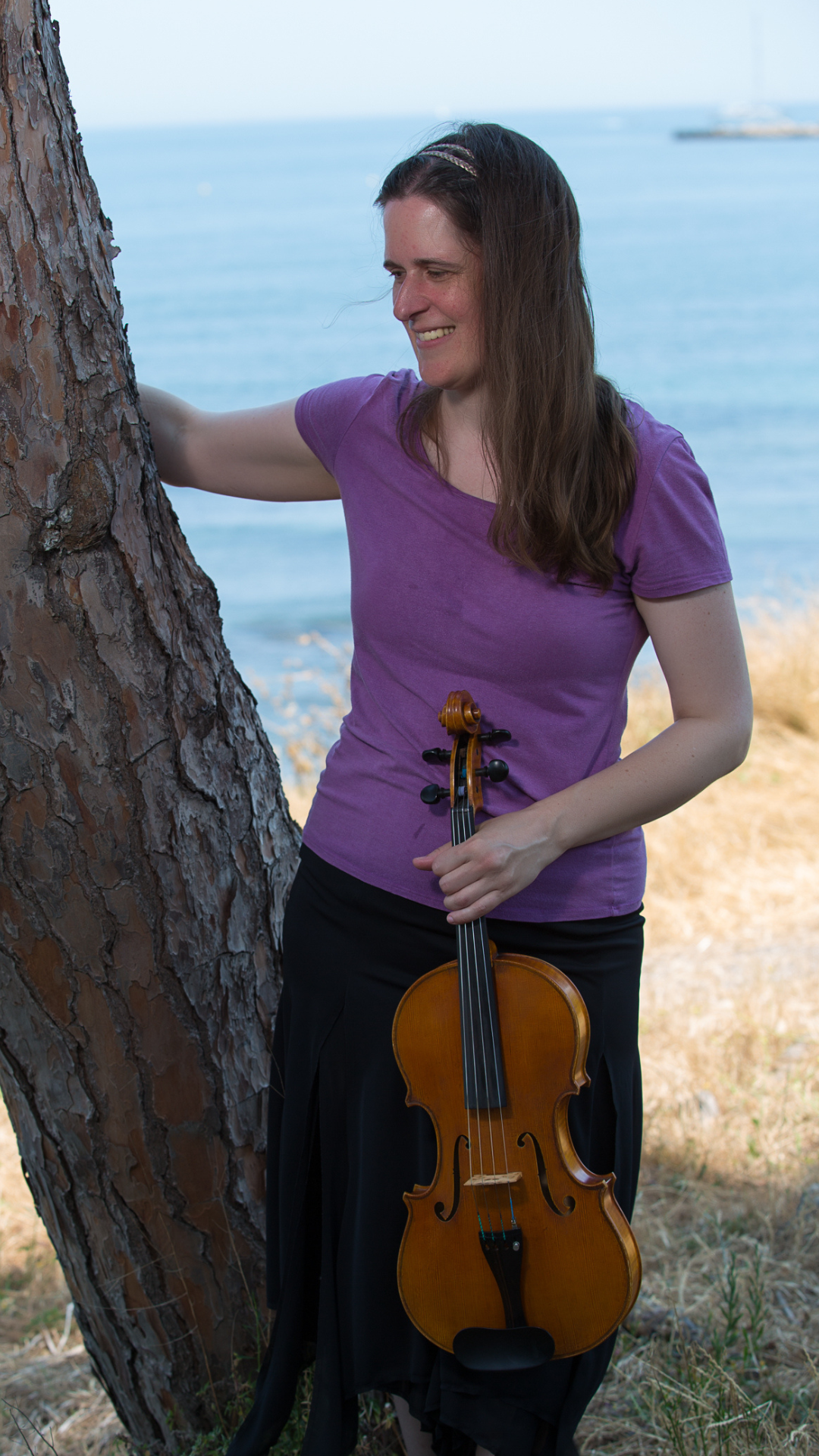 Leah Irby in a purple shirt and black skirt standing with a viola in her hands next to a tree overlooking the water.
