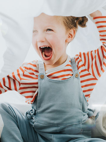 Child in an excited mouth open posture wearing a red striped shirt and jeans overalls