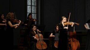 An all female chamber orchestra, you can see a violinist playing solo and other cello, violin and viola players surrounding her with the window of a church behind them.