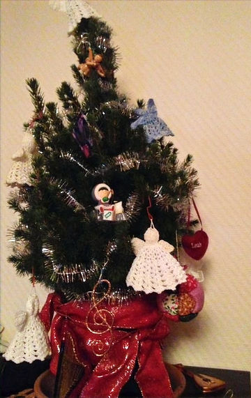 Small tabletop christmas tree decorated with a few crocheted angels and other ornaments and a red bow around the bottom of the pot.