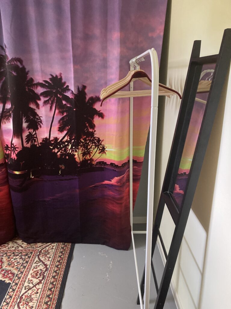 room with a sunset curtain and persian rug on the floor and a standing mirror that is framed in black and shows the sunset reflected in its surface.